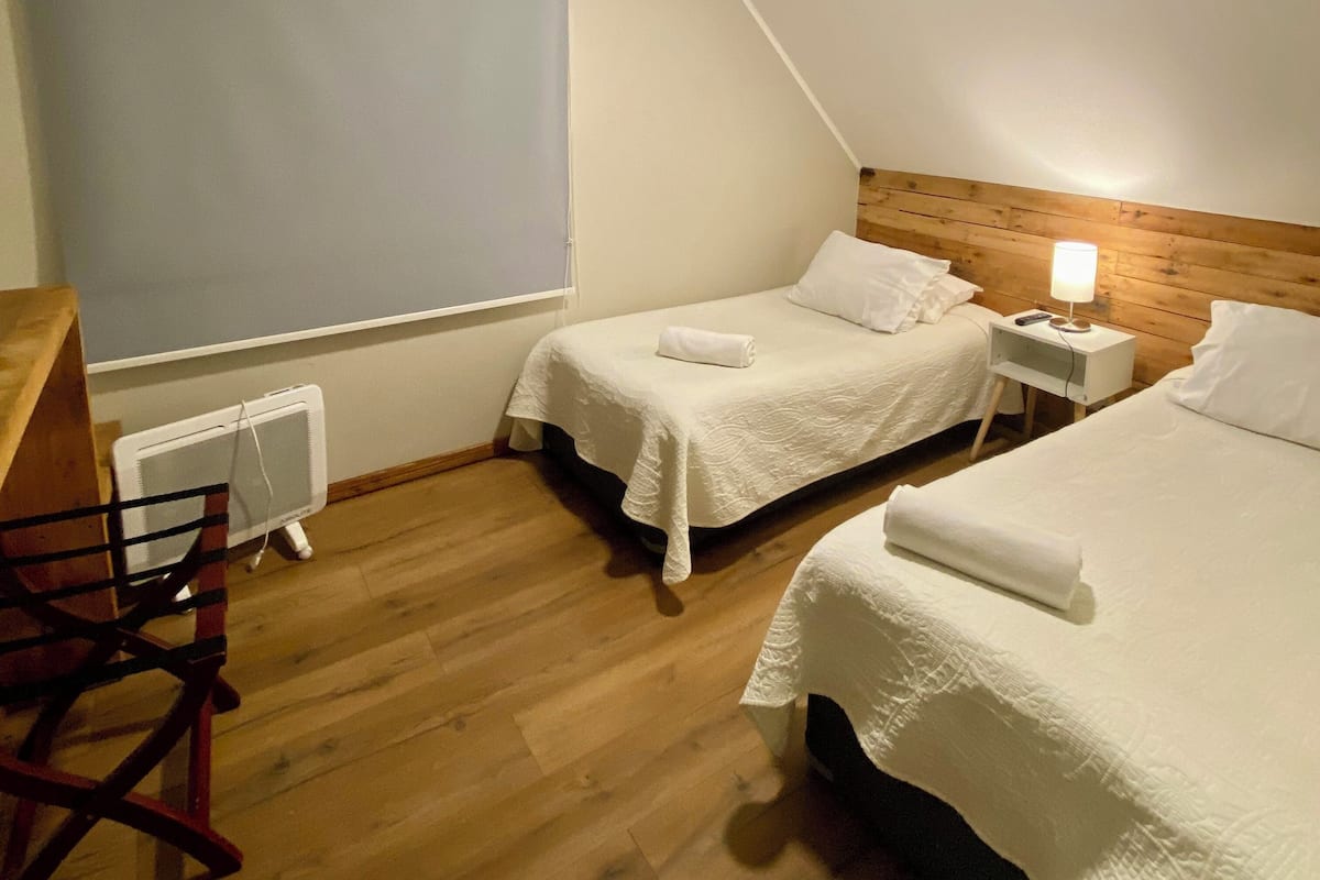 A room with two beds, a desk, and a chair. There is a window with a blind, a heater, and a lamp on the nightstand.