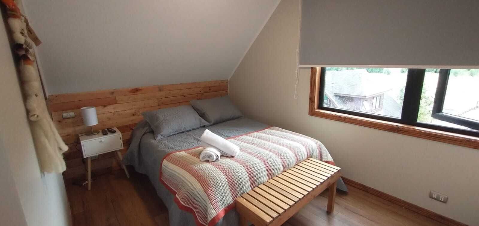 A bedroom with a sloped ceiling, a wooden headboard, a bed with a striped blanket, a bedside table with a lamp, and a window with a view of the outside.