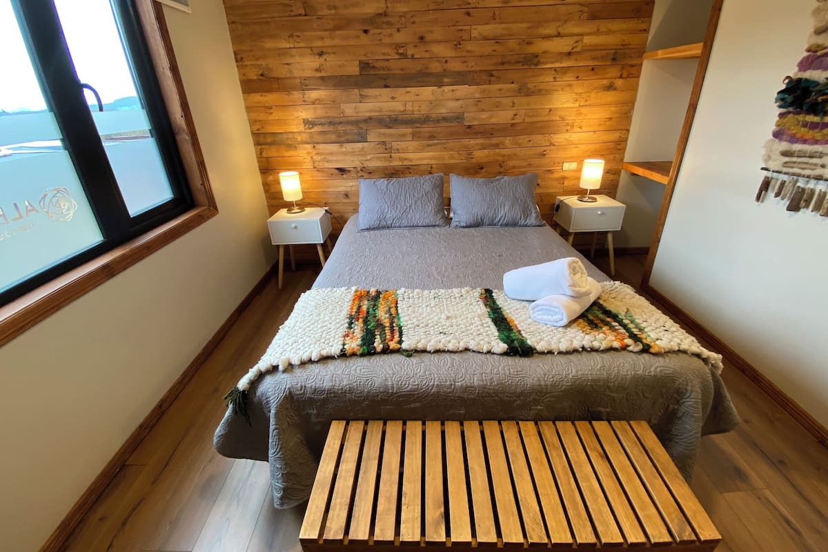A hotel room with a wooden headboard, a large bed with a gray bedspread, two bedside tables with lamps, and a view of the sea through a window.