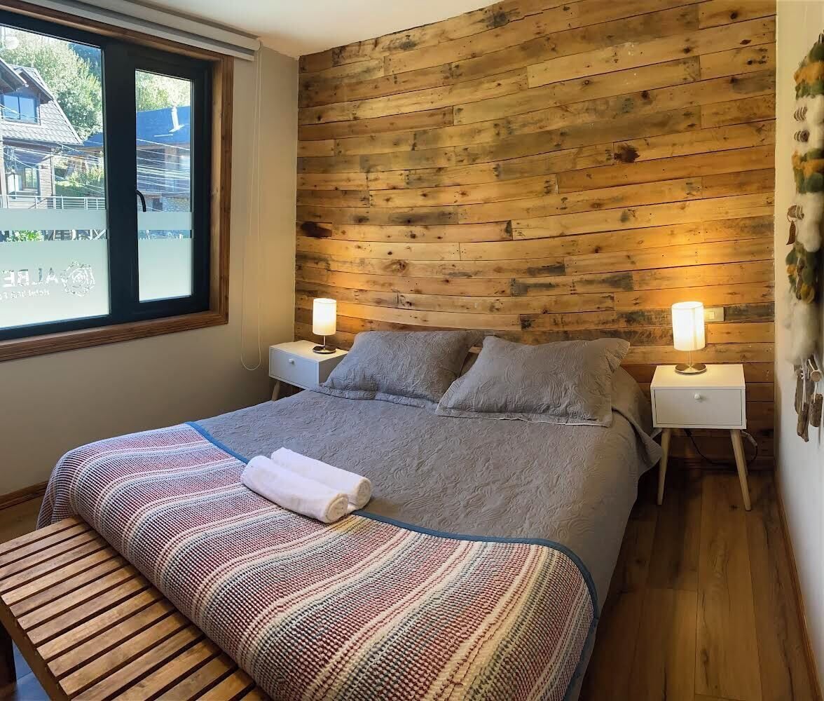 A bedroom with a wooden headboard, a bed with grey bedding, two bedside lamps, a bench, and a window with a view of buildings.
