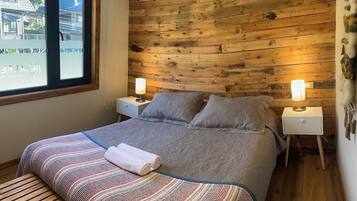 A bedroom with a wooden headboard, a bed with grey bedding, two bedside lamps, a bench, and a window with a view of buildings.