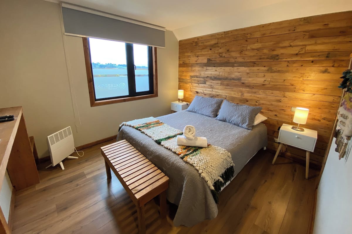 A bedroom with a wooden headboard, a large window with a view of a body of water, a bed with a blanket, pillows, and a bedside lamp.