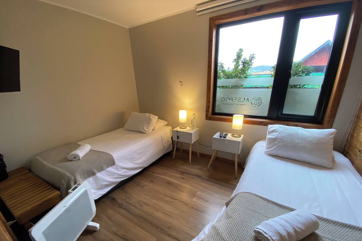 A hotel room with two single beds, a wooden floor, a radiator, a small table with a lamp, and a window with a view of greenery and a building.