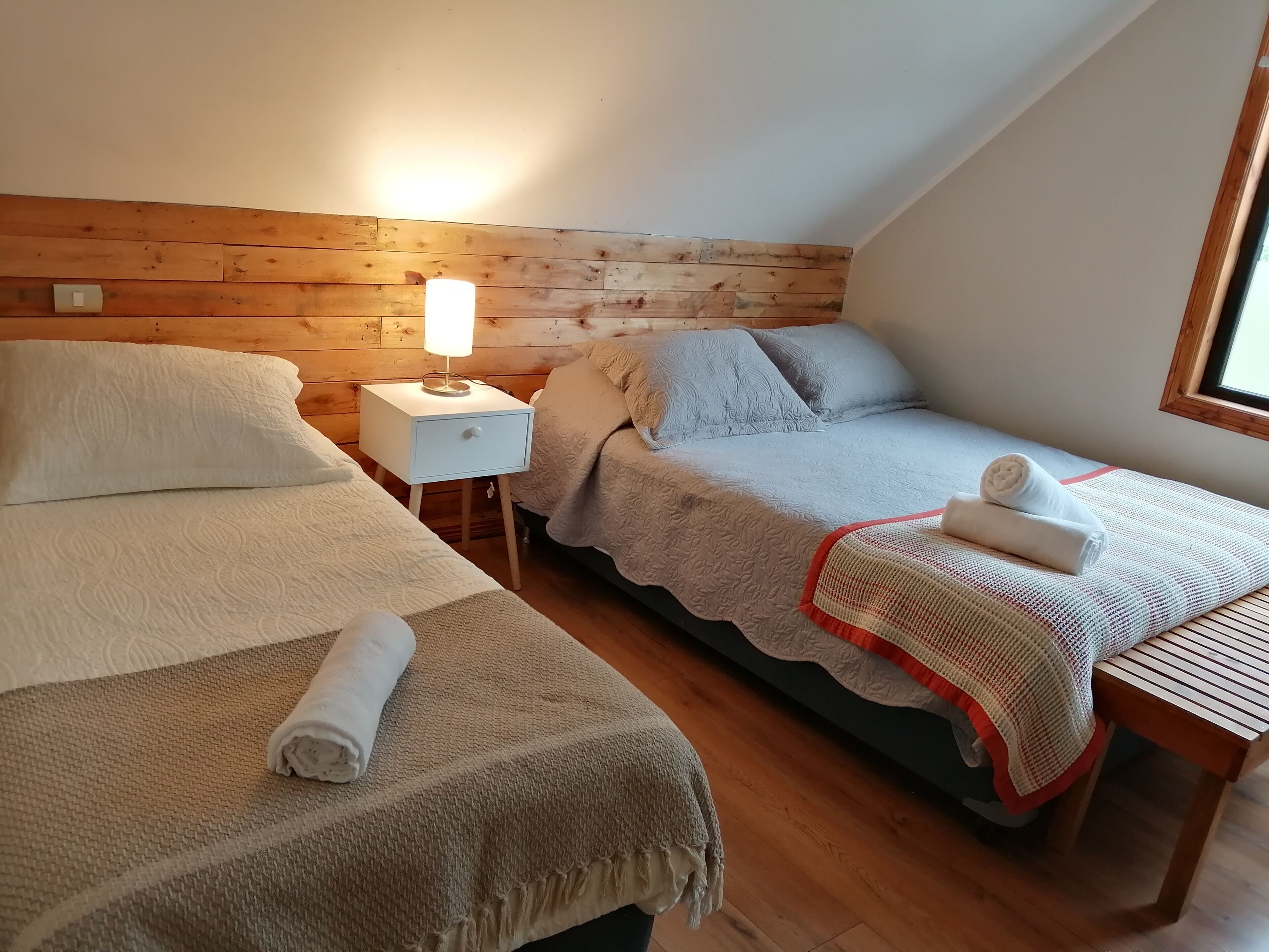 A cozy bedroom with two beds, a nightstand with a lamp, and a window providing natural light.