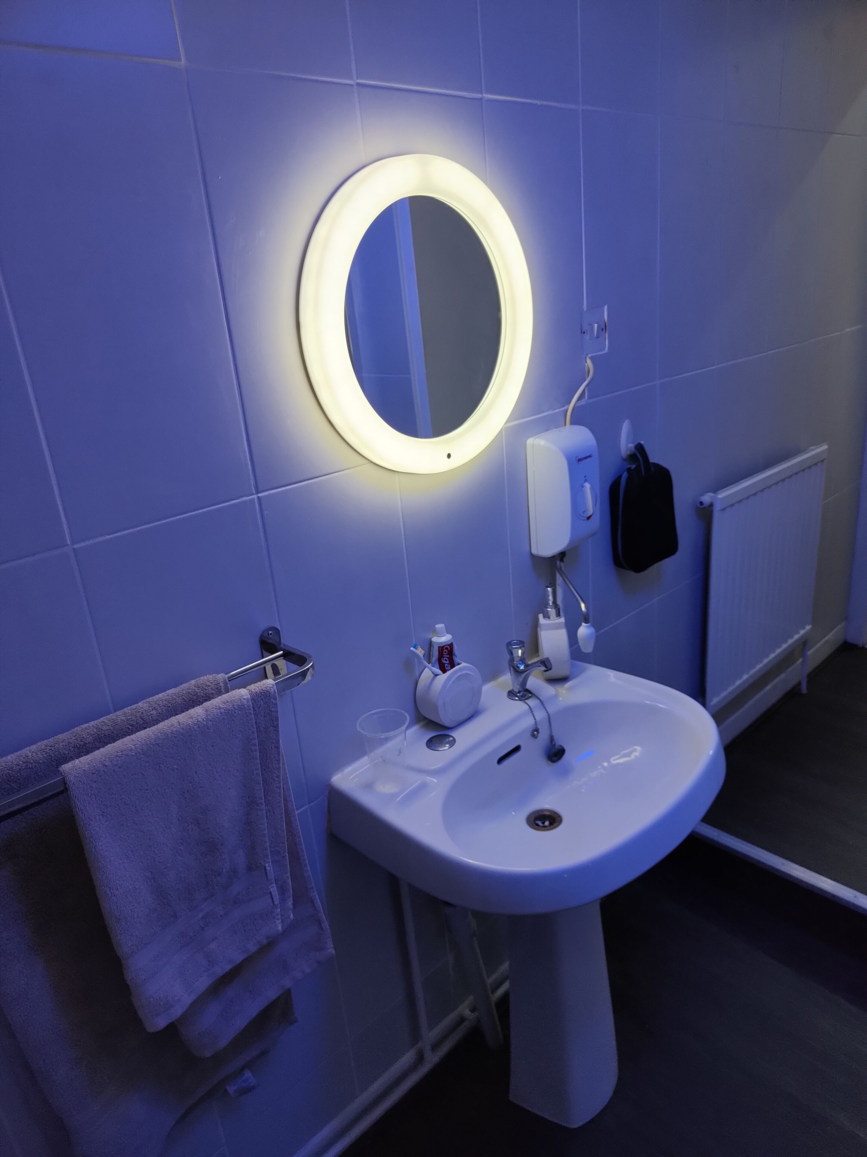 Een badkamer met een wit wastafelmeubel, een ronde spiegel met verlichting, een handdoekrek met handdoeken, een zeepdispenser en een zwarte tas aan de muur.