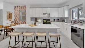 Una cocina moderna con armarios blancos, una isla central con taburetes y un lavavajillas de acero inoxidable.