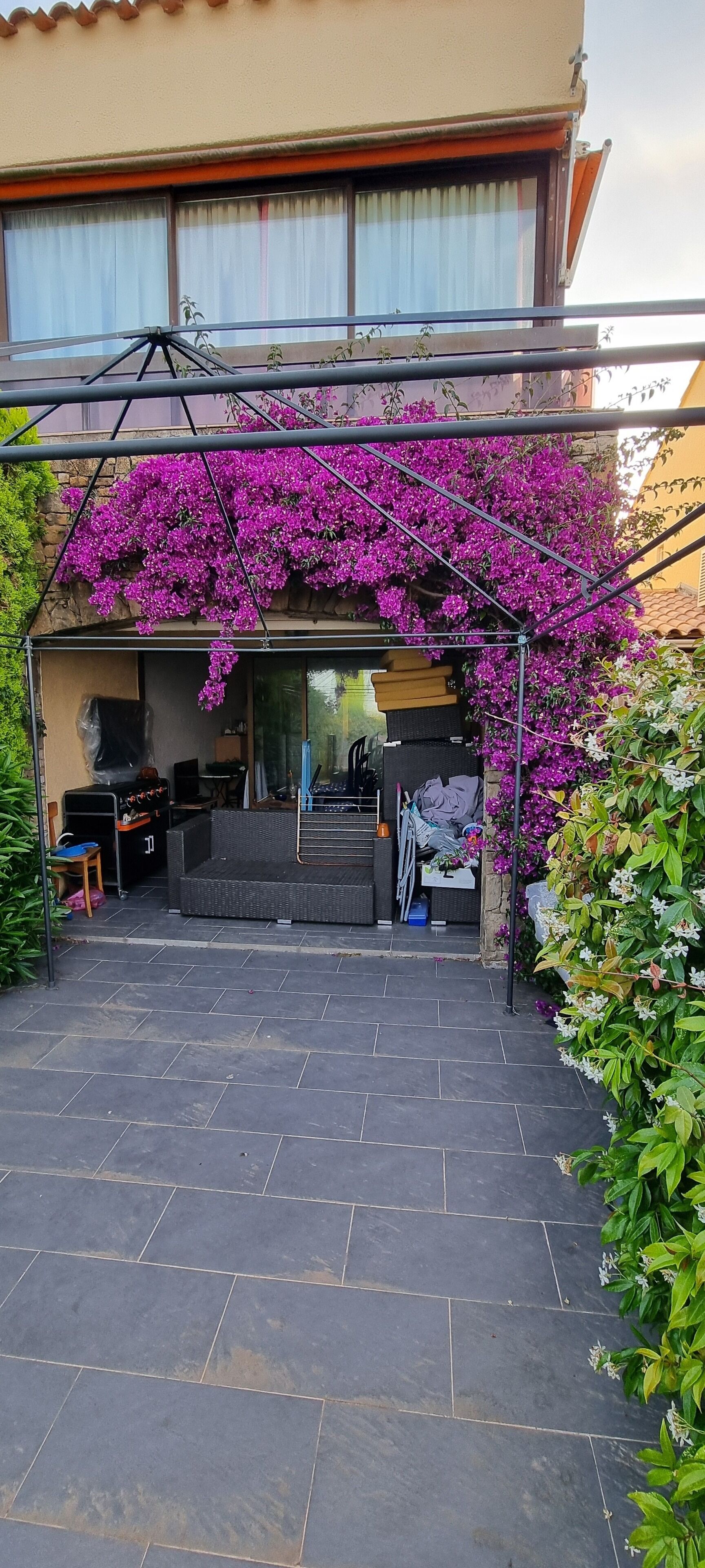 Een overdekt terras met stenen vloer, een zithoek met kussens en een pergola met paarse bloeiende planten.