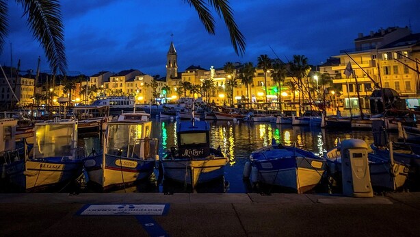 Un port de plaisance au crépuscule, avec des bateaux amarrés, une tour imposante et des bâtiments illuminés d’une lumière chaleureuse.