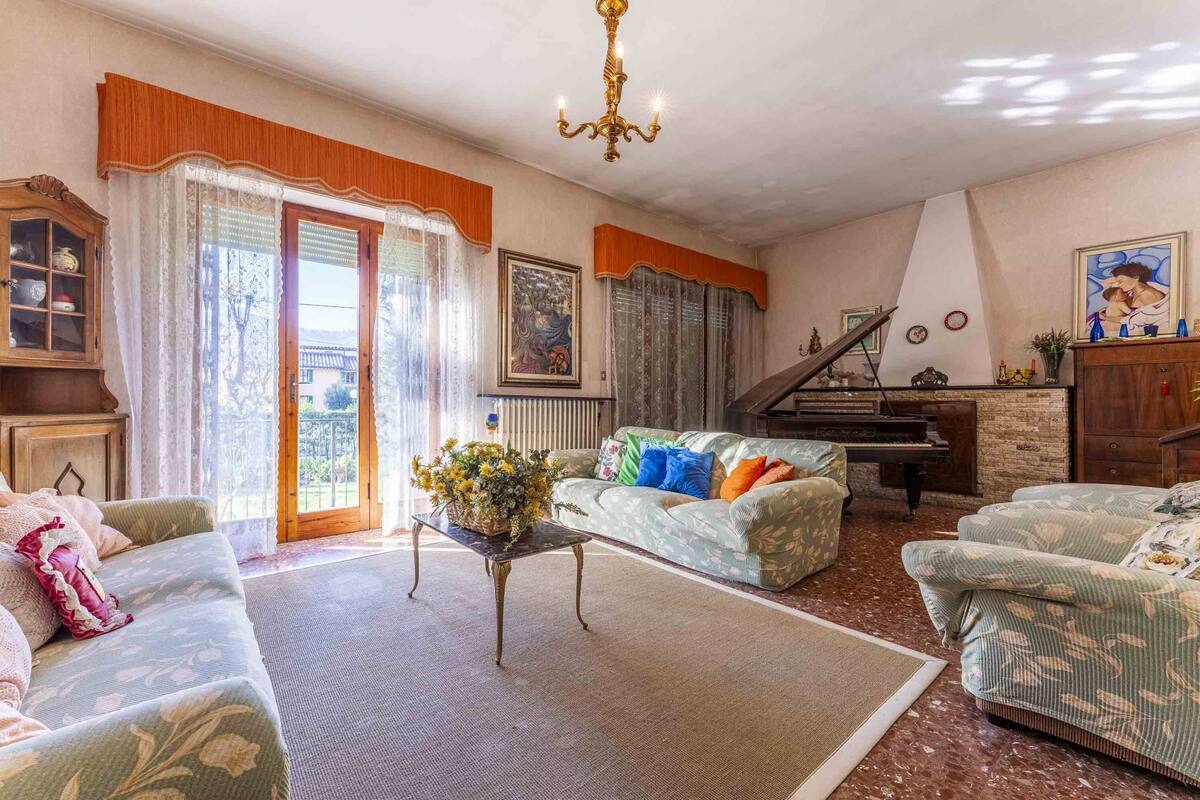 A cozy living room with a piano, a sofa, a coffee table with flowers, and a large window with curtains.