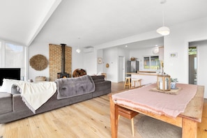 Ein modernes Wohnzimmer mit einem grauen Sofa, einem Holzesstisch mit rosa Tischläufer, einem Kamin und einer Küche im Hintergrund.