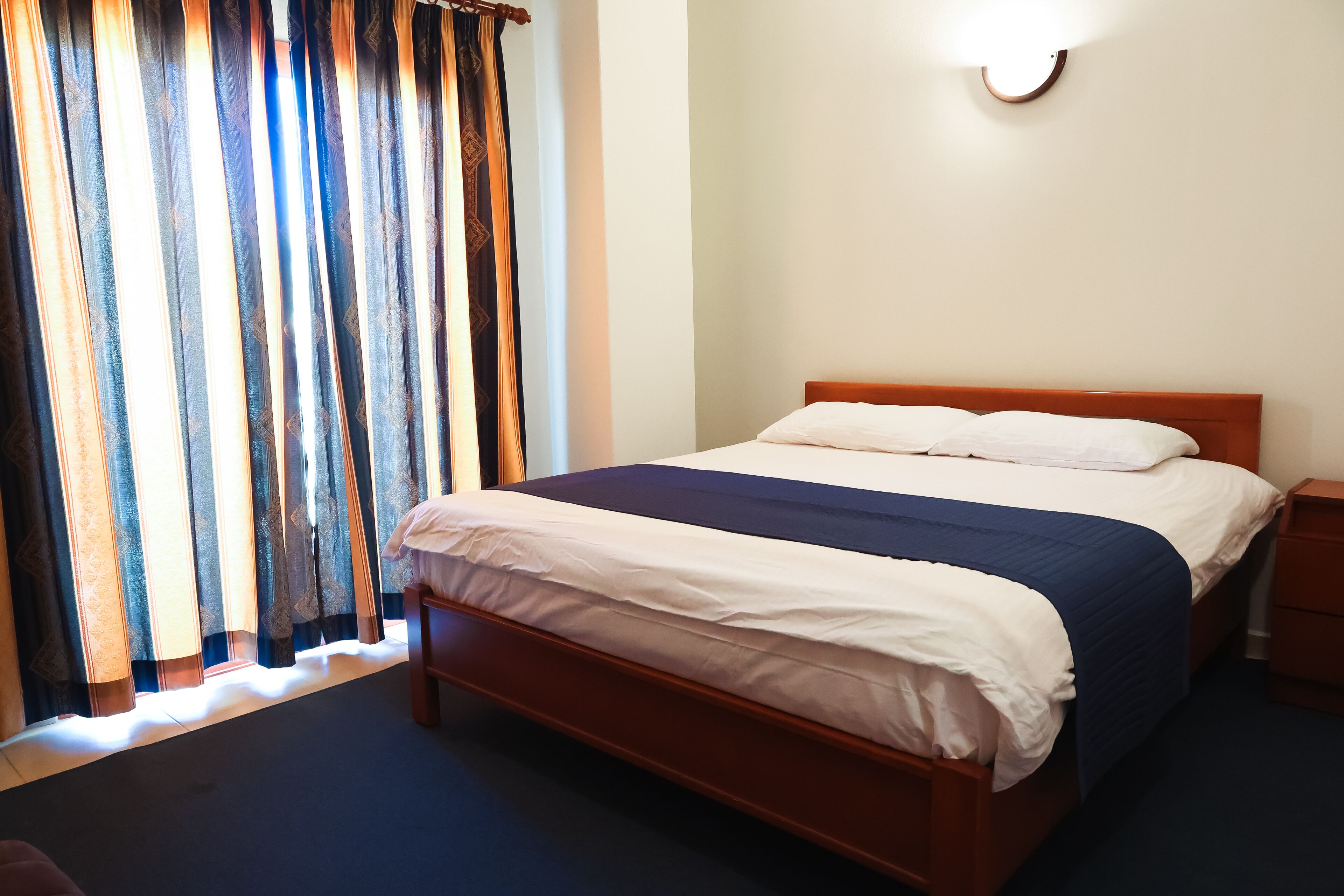 A neatly made bed with white and blue bedding, a wooden headboard, and a bedside table with a lamp.