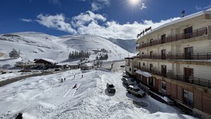 Un pueblo nevado al pie de una montaña, con un edificio que dice "Ecole du Meilleur" y balcones, y una pista de esquí en el fondo.