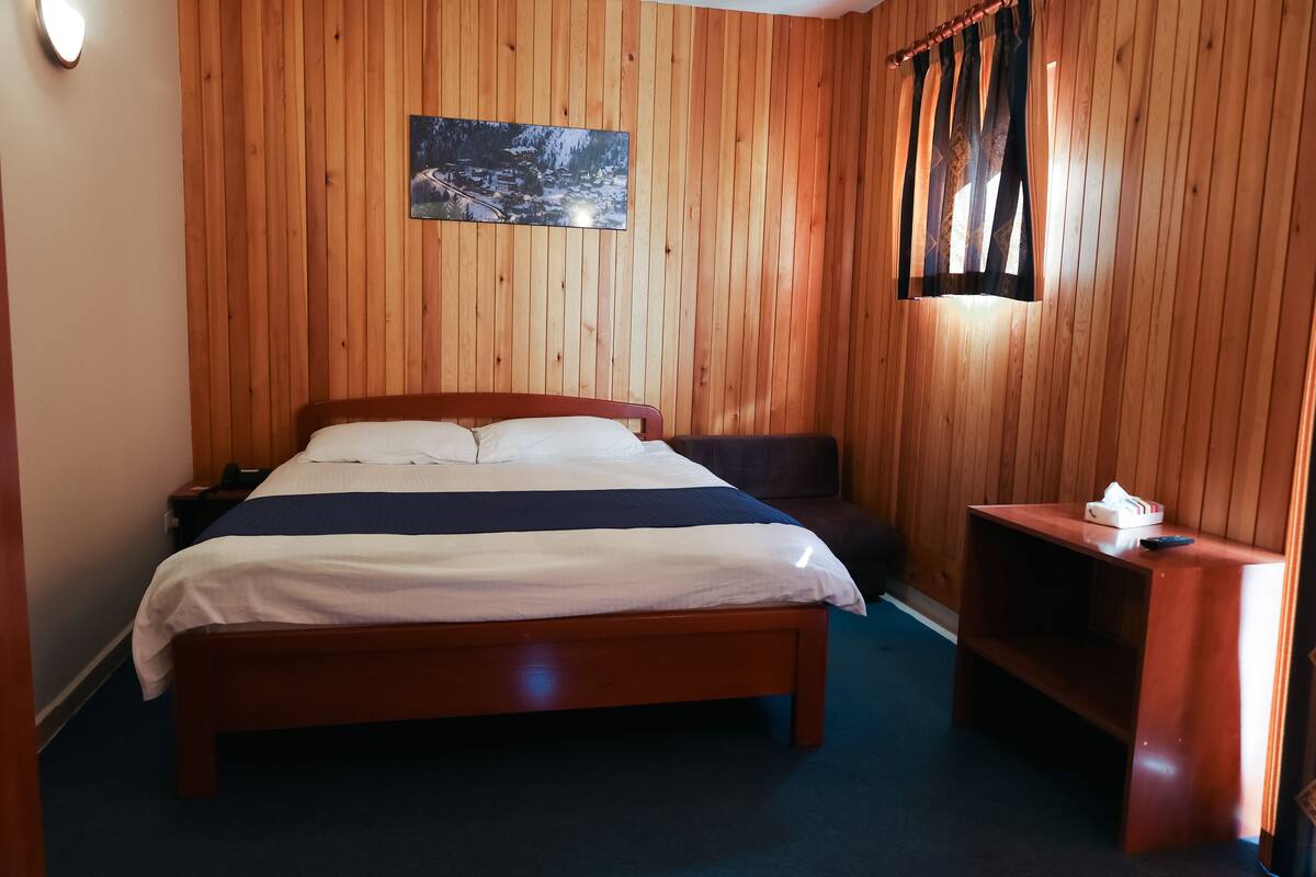 A wooden-paneled room with a bed, a nightstand, a small table, and a painting on the wall.