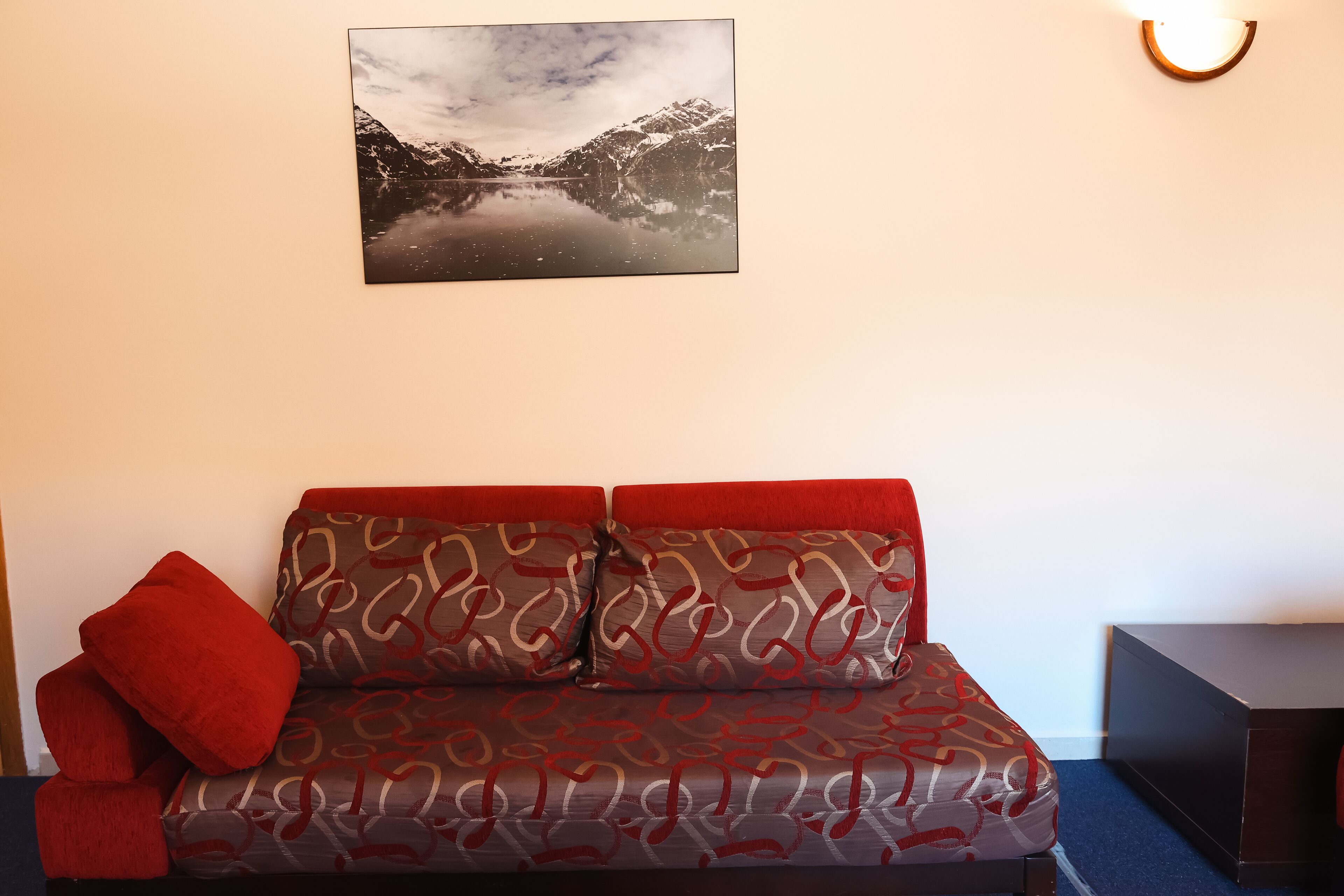 A room with a sofa, a red pillow, a wall-mounted picture of a mountain landscape, and a small table with a dark surface.