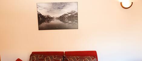 A room with a sofa, a red pillow, a wall-mounted picture of a mountain landscape, and a small table with a dark surface.