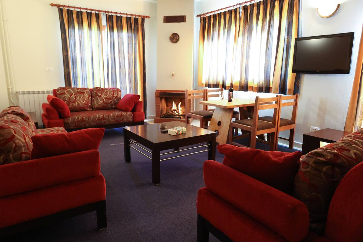 A living room with a fireplace, red sofas, a wooden dining table, and a television mounted on the wall.