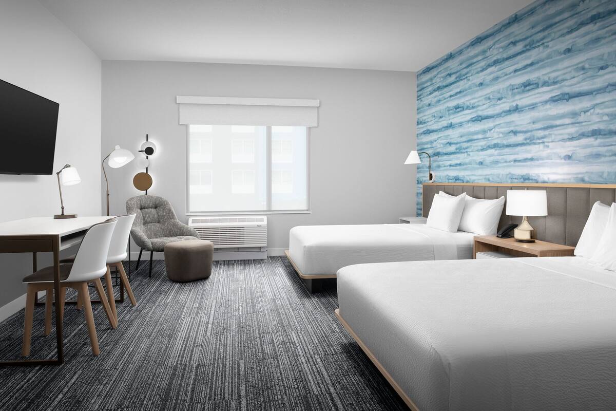 Une chambre d’hôtel avec un grand lit, un lit plus petit, un bureau avec des chaises, une lampe de bureau, une chaise, une table de chevet avec une lampe et une grande fenêtre avec des stores.