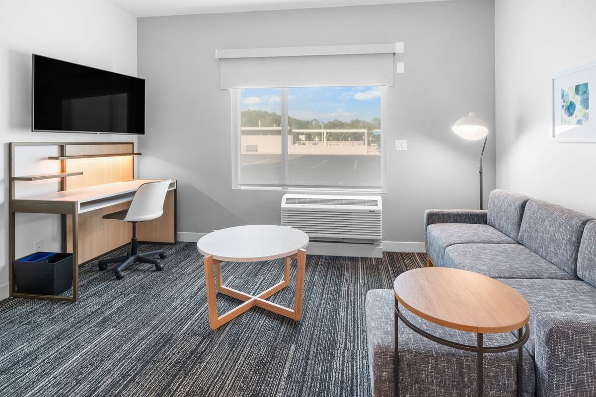 Une chambre d’hôtel moderne équipée d’un bureau, d’un canapé, d’une table ronde, d’une télévision et d’une grande fenêtre donnant sur l’extérieur.