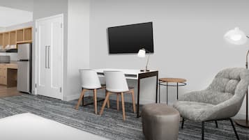 A modern kitchen with white cabinets, a refrigerator, and a microwave. There is a dining area with a table, chairs, and a lamp.