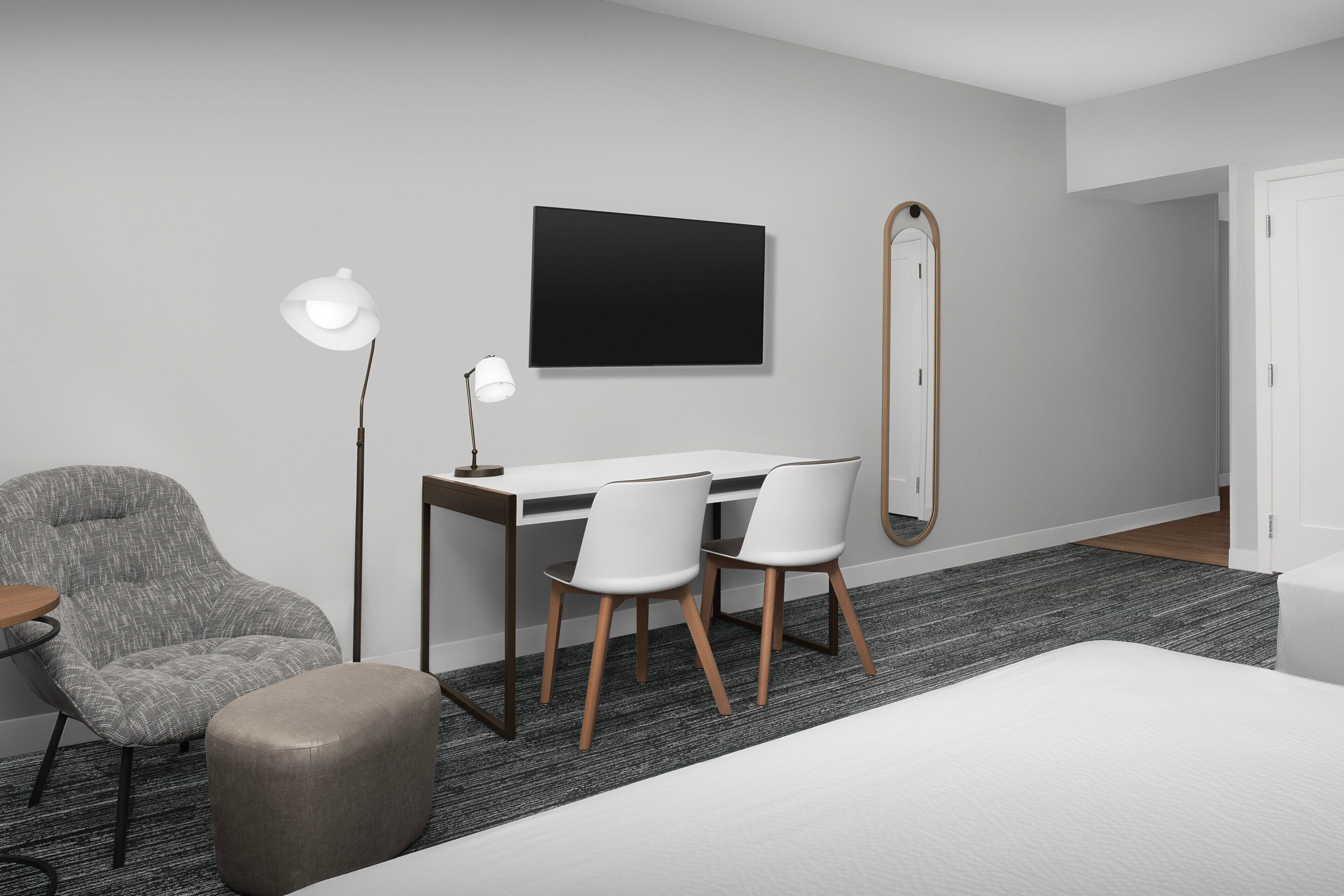 Una habitación de hotel moderna con escritorio, dos sillas, un espejo grande, una lámpara y una cama.
