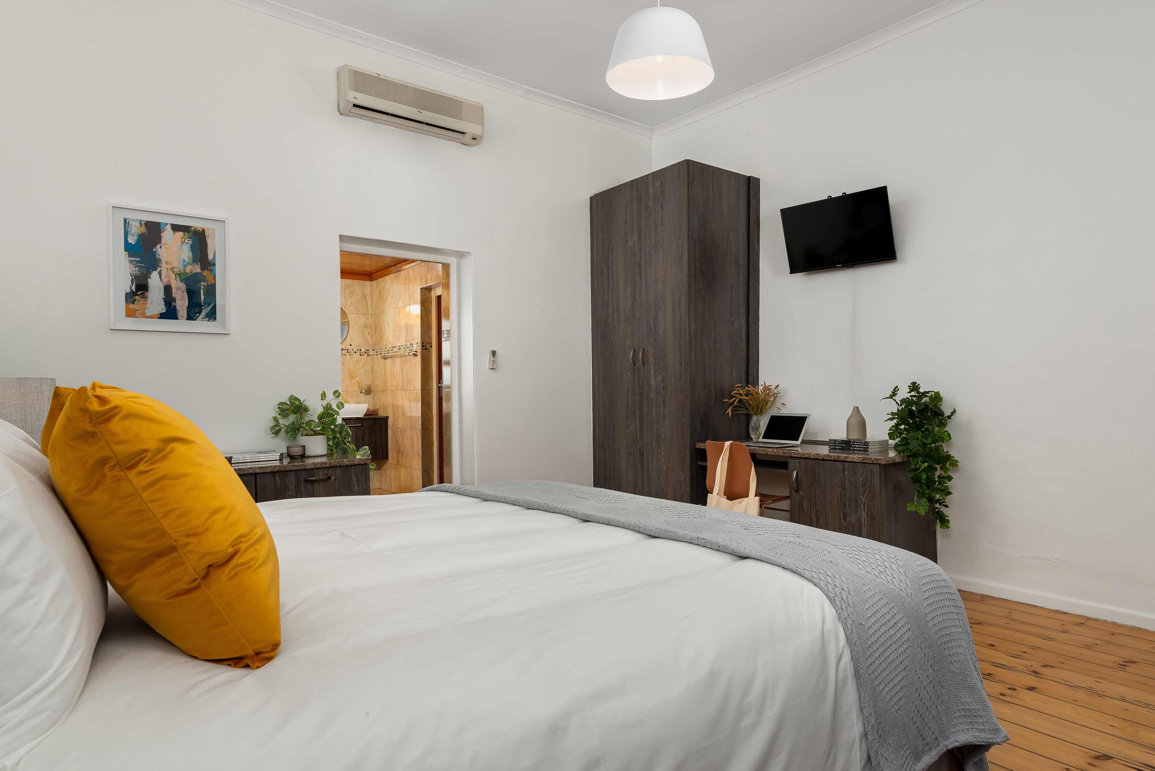 A modern bedroom with a large bed, a wardrobe, a desk with a plant, and a TV on the wall.