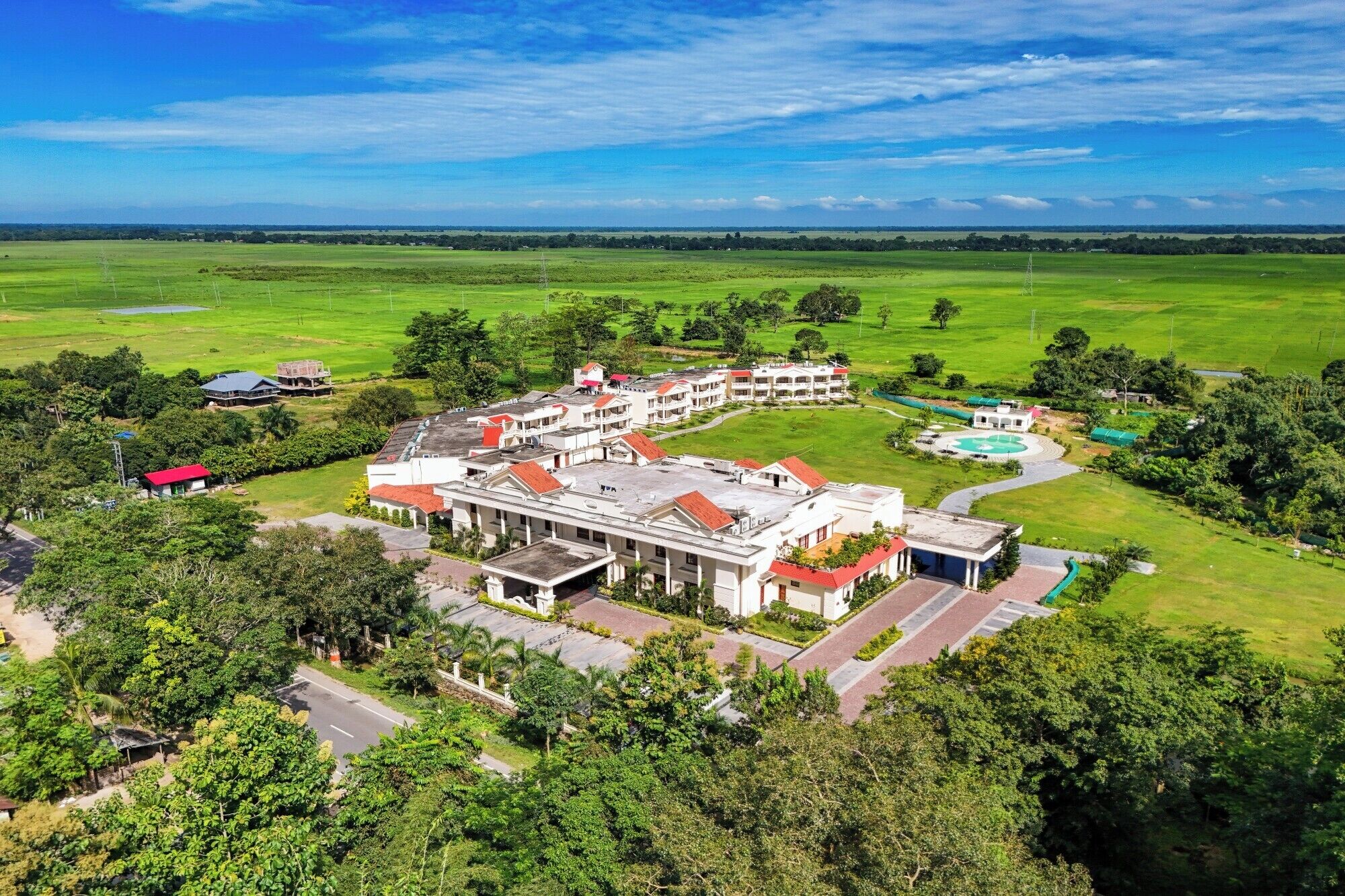 An aerial view of a hotel with multiple buildings, a swimming pool, and surrounding green fields.