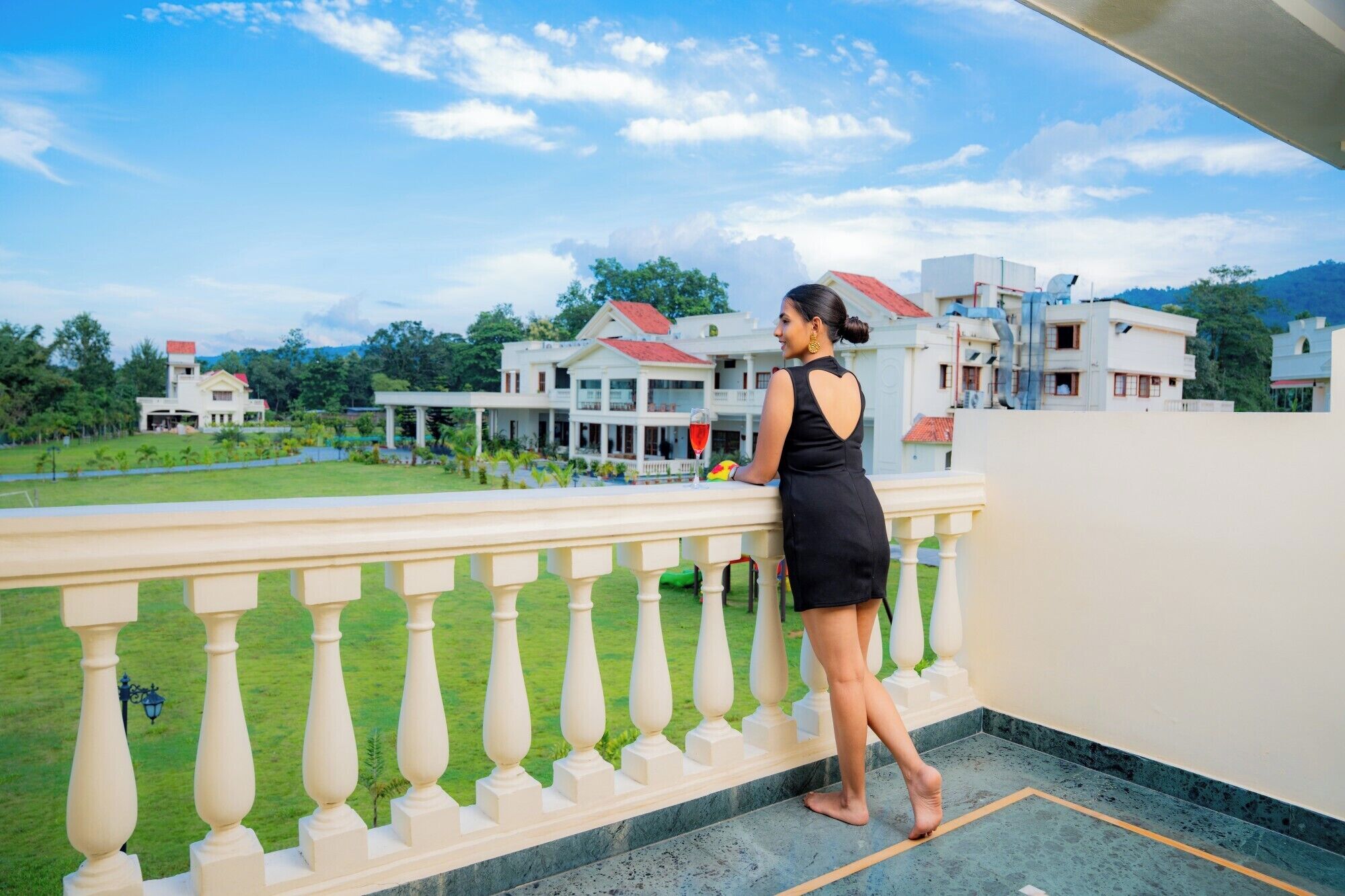 A woman standing on a balcony with a view of a large building complex and a green lawn.