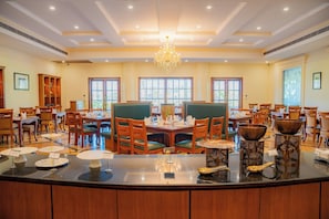 A spacious dining area with multiple tables and chairs, a central chandelier, and large windows allowing natural light.