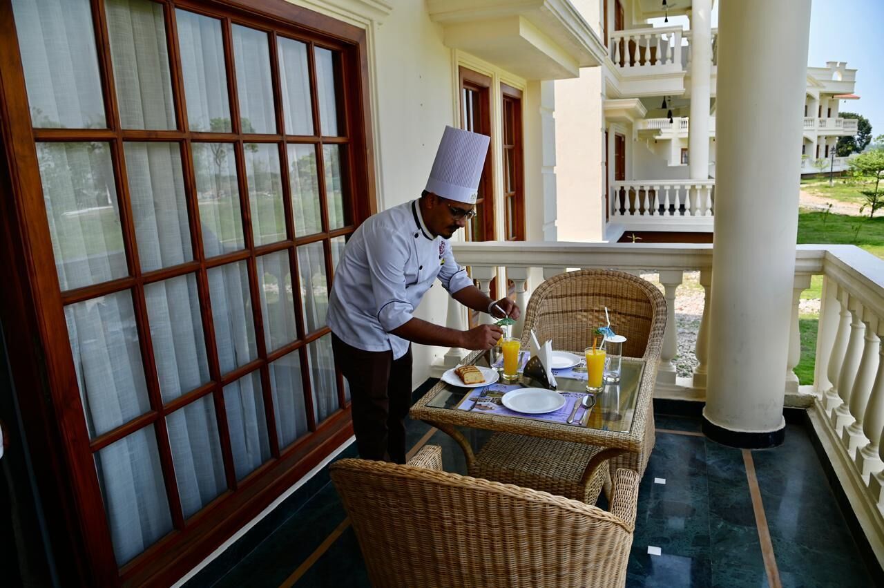 A chef preparing breakfast on a balcony with wicker furniture, a glass table, and a view of the building's exterior.