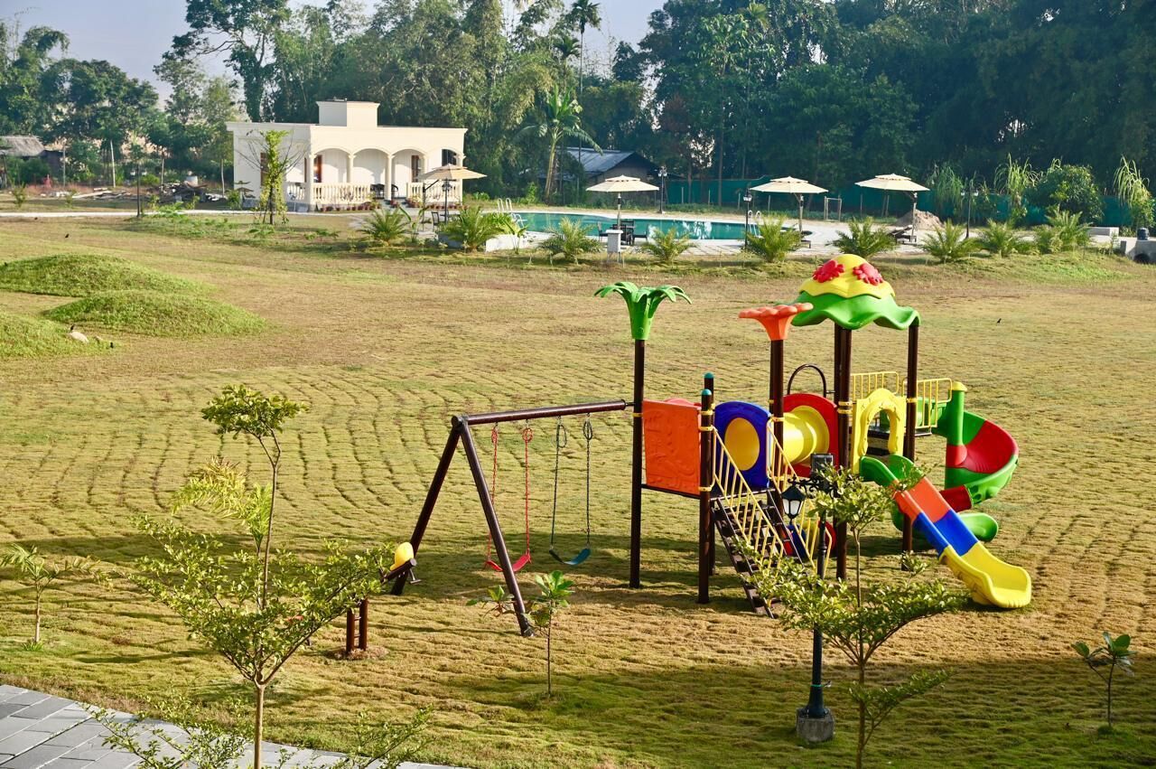 A playground with swings and slides, surrounded by a grassy area and a pool in the background.