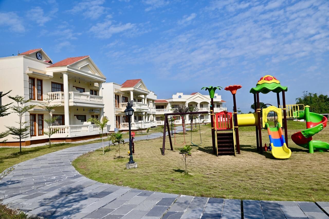 A playground with slides and climbing structures, a paved pathway, and a building with balconies and a red roof.