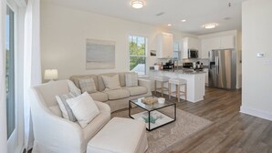Una sala moderna con un sofá color crema, una mesa de centro de vidrio y una cocina con electrodomésticos de acero inoxidable.