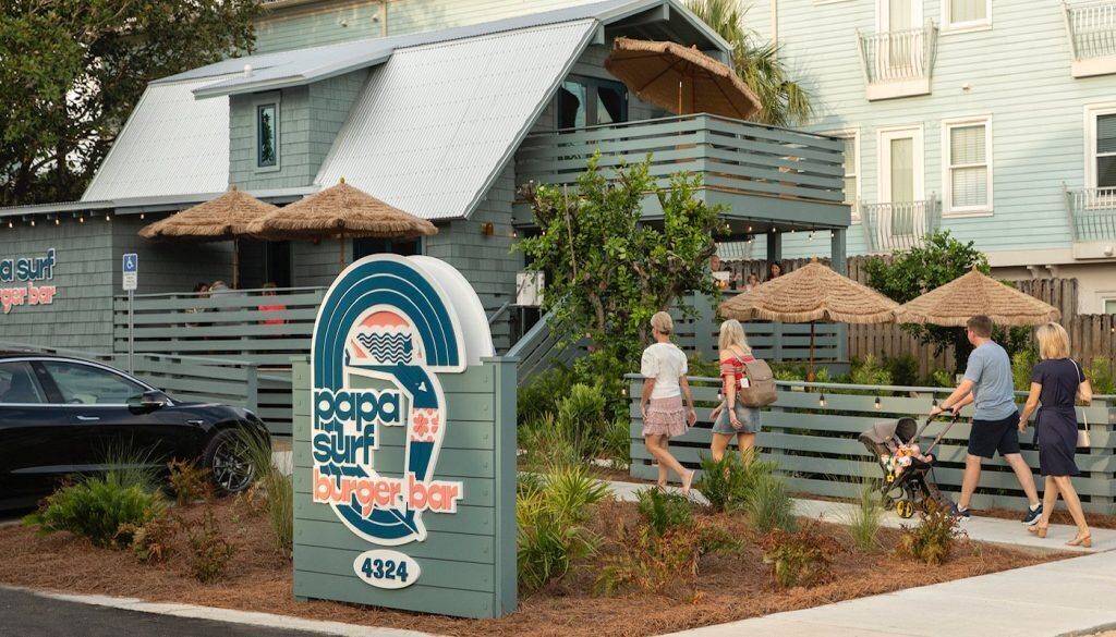 Un restaurant amb temàtica de platja, un cartell amb una tabla de surf, zones d'aixopluc a l'exterior i una tanca de fusta.