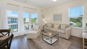 Amplia sala de estar con sofá, sillón y mesa de centro, ventanas grandes con persianas, y un balcón con vistas a edificios y zonas verdes.
