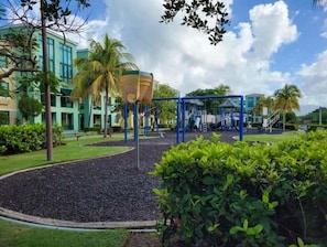 Children can play on the swing set and climbing structure amidst lush greenery and palm trees.
