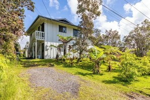 Une maison à deux étages dotée d’un balcon, entourée d’une végétation luxuriante et bordée d’un chemin de gravier.