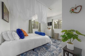 A bedroom with a bed, pillows, a plant, a TV, and a window.