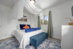 Une chambre Ă coucher avec un lit, un banc, une fenĂȘtre avec des rideaux et un tableau au mur.
