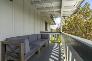 A covered porch with a gray sofa and wooden frame, white walls, and a view of trees.