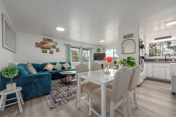 A modern living room with a teal sofa, a dining table, and a kitchen area with white cabinets.