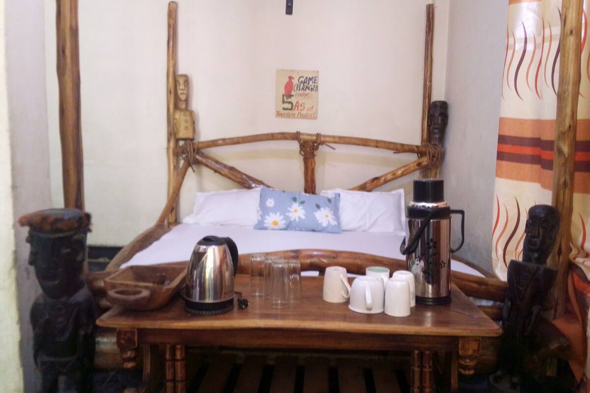 A wooden bed with a woven headboard, a small table with a kettle, cups, and a bottle.
