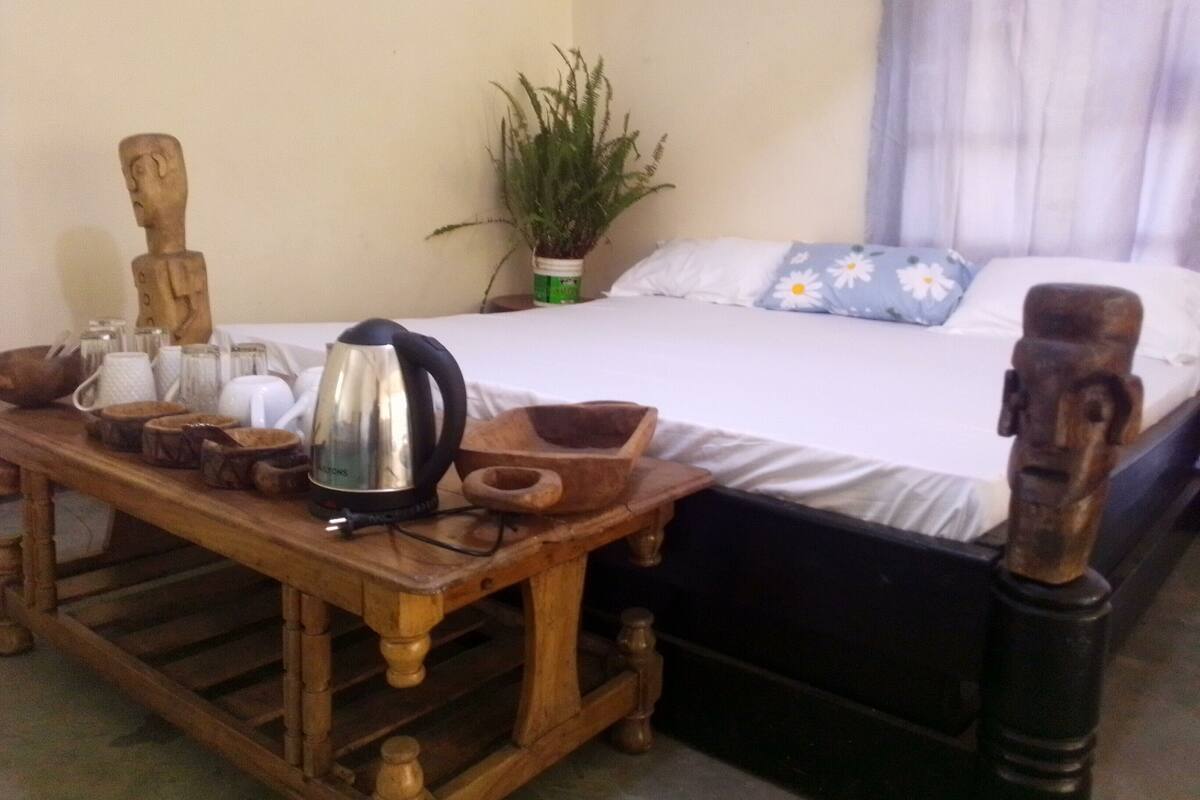 A wooden table with a kettle, cups, and bowls, a bed with a white sheet and a blue pillow, and a green plant in the corner.
