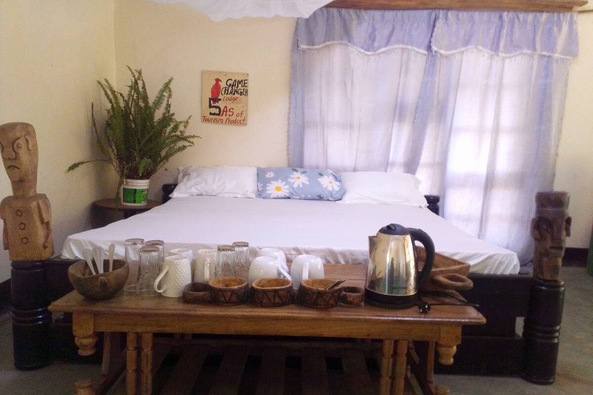 A neatly made bed with a white bedspread and blue floral pillows, a wooden table with various items like a kettle and glasses, and a sign on the wall advertising a game-changing lodge.