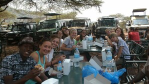 Un grupo de personas sentadas en una mesa al aire libre, con cajas de comida y botellas de agua.