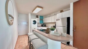 A compact kitchen with a washing machine, a refrigerator, and a sink. There is a bar counter with stools and a potted plant.