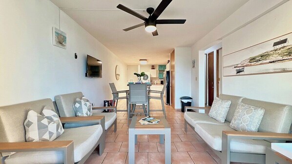 A living room with two sofas, a coffee table, and a ceiling fan. There is a dining area with chairs and a table in the background.