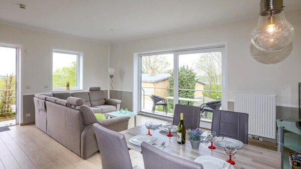 A modern living room with a grey sofa, a dining table set for four, and large windows offering a view of the outdoors. - Amselresidenz 4 - STAR (Binz)