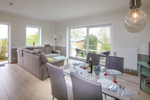 A modern living room with a grey sofa, a dining table set for four, and large windows offering a view of the outdoors.