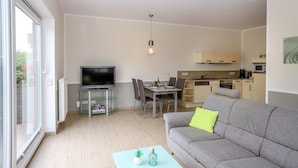 A modern living room with a grey sofa, a TV on a glass stand, a dining area with a table and chairs, and a kitchen in the background. - Amselresidenz 4 - STAR (Binz)