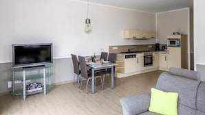 A modern living room with a flat-screen TV, a glass TV stand, a dining table with chairs, a grey sofa, and a kitchen area with cabinets and appliances. - Amselresidenz 4 - STAR (Binz)